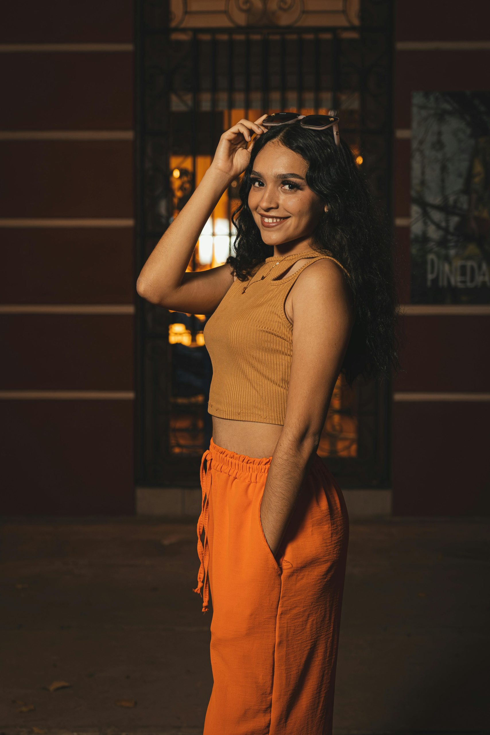 Confident woman with long black hair smiling indoors, wearing sunglasses and vibrant outfit.